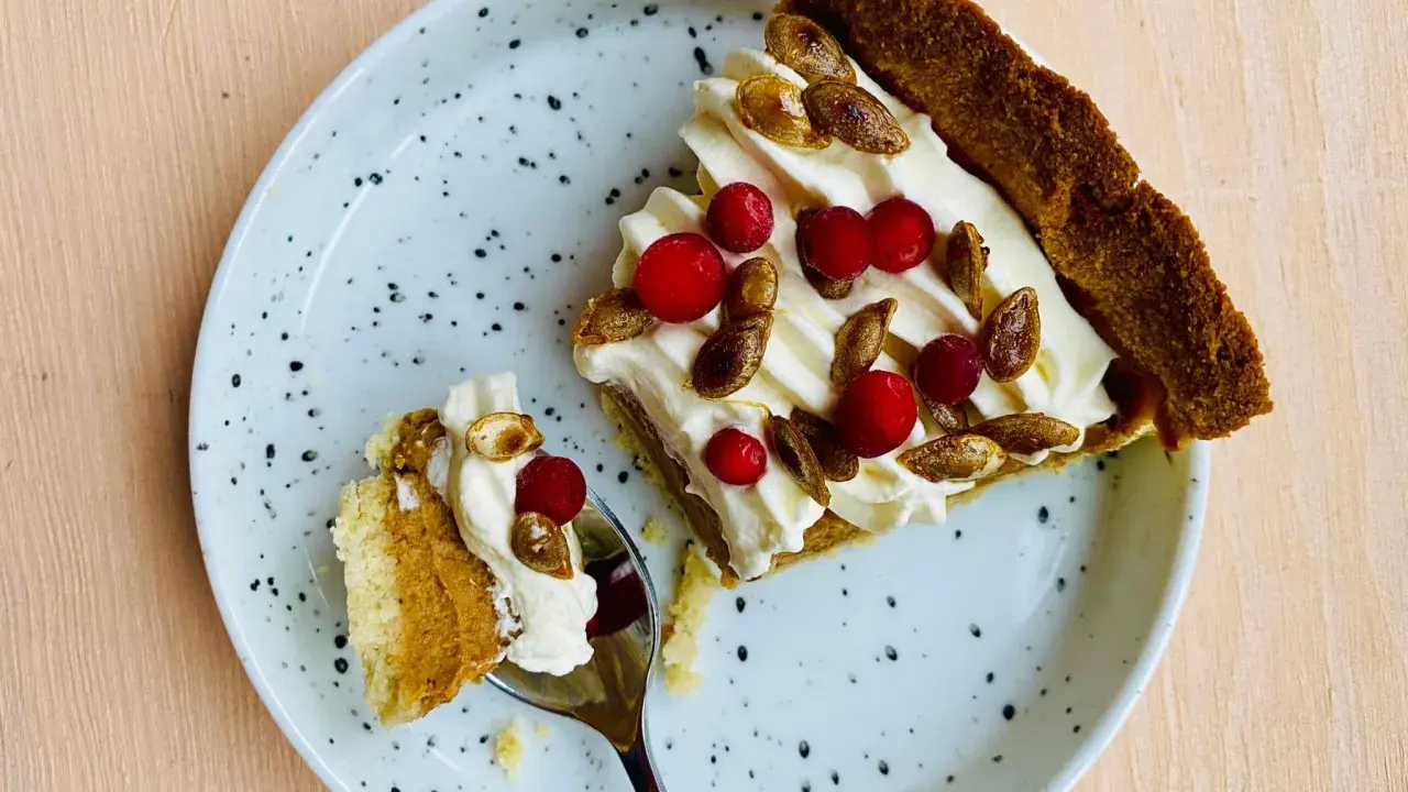 En bit glutenfri pumpapaj med vispgrädde, lingon och rostade pumpafrön på en prickig tallrik. En sked med en bit paj ligger bredvid.