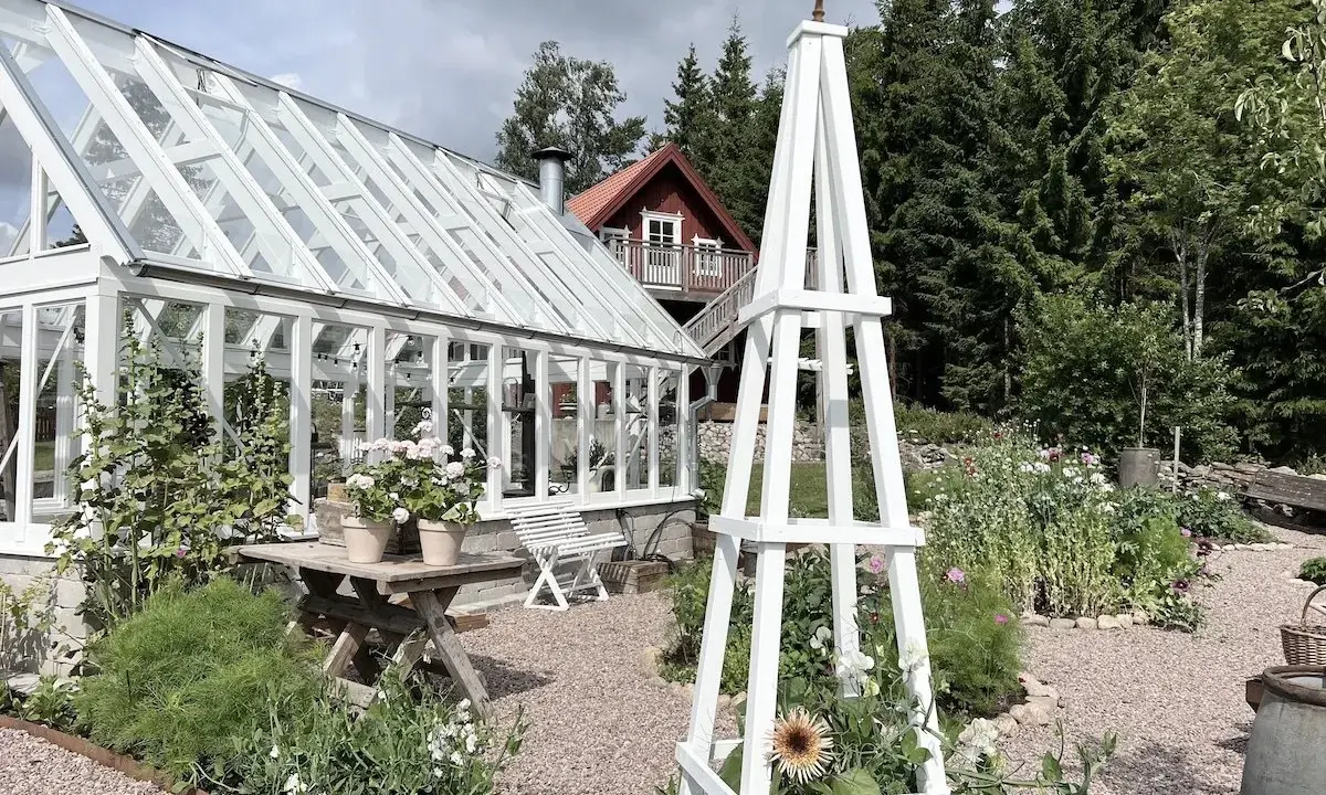 En vit rosentorn ritning står i en lummig trädgård med växthus och blommor.
