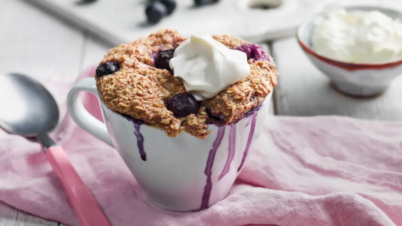 En nyttig mugcake med blåbär och en klick grädde, serverad i en vit mugg med lila droppar. En sked och en skål med grädde syns i bakgrunden.