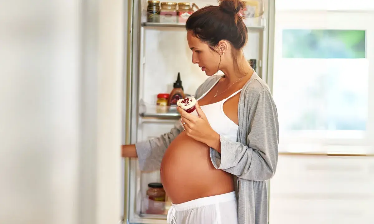Gravid kvinna med stor mage tar en cupcake ur kylskåpet, kanske för att stilla sina cravings.