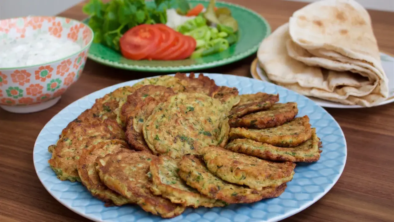 En stor tallrik med gyllene zeinas zucchinibiffar, serverade med pitabröd, en skål med vit sås och en sallad med tomat och gurka.