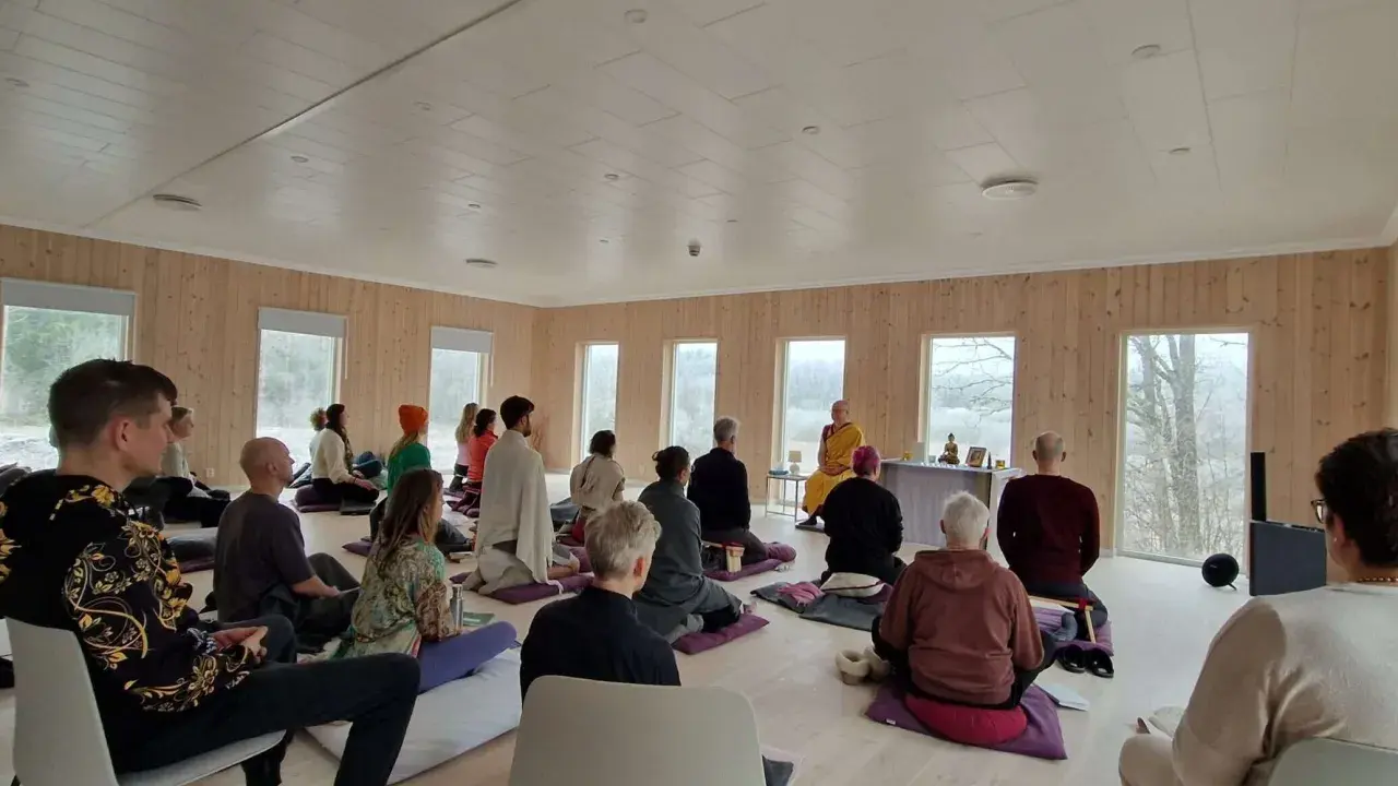 Människor sitter på kuddar och stolar under en meditation retreat i Sverige. En person i munkkläder leder sessionen.