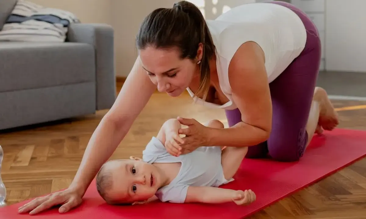 En mamma på knä på en röd yogamatta leker med sin bebis. Hon hjälper till med bebisens ben. En fin stund för post partum-träning.