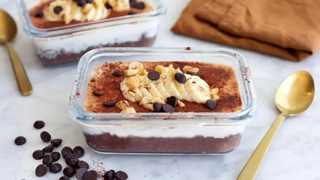 Två glasbehållare med nyttig chokladpudding, toppad med banan, nötter och chokladknappar. En gyllene sked ligger bredvid.