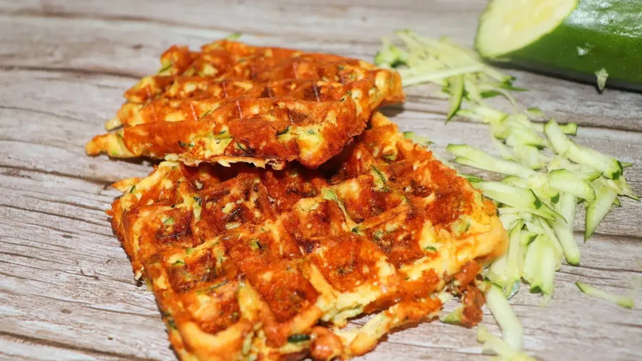 Gyldene zucchini våfflor, perfekt stekta, med riven zucchini och en hel zucchini i bakgrunden.
