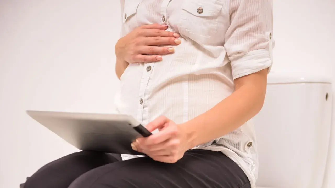 Gravid kvinna sitter på toaletten med en surfplatta, oroar sig för brun flytning.