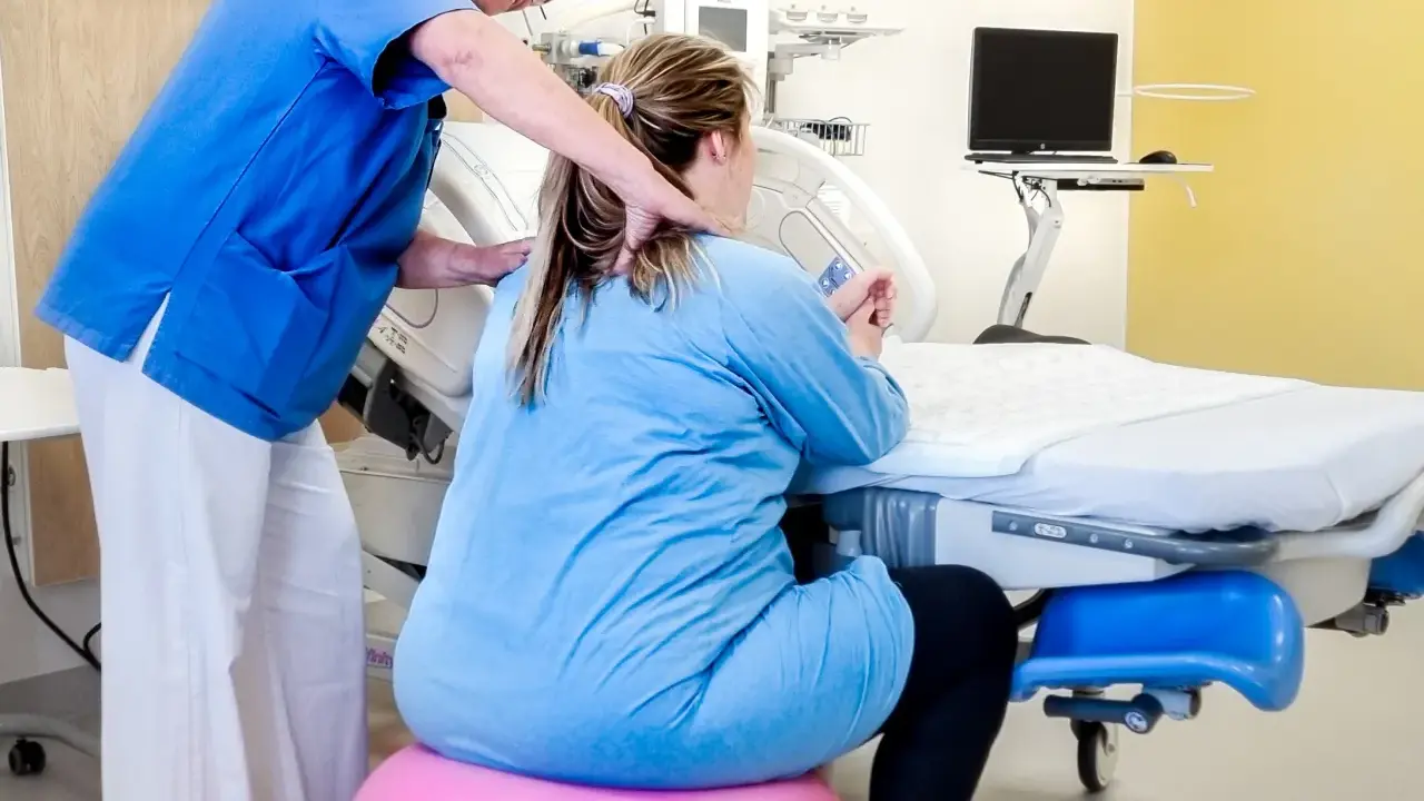 Barnmorska ger stöd till en kvinna i gravid vecka 36 som sitter på en rosa boll vid en förlossningssäng.