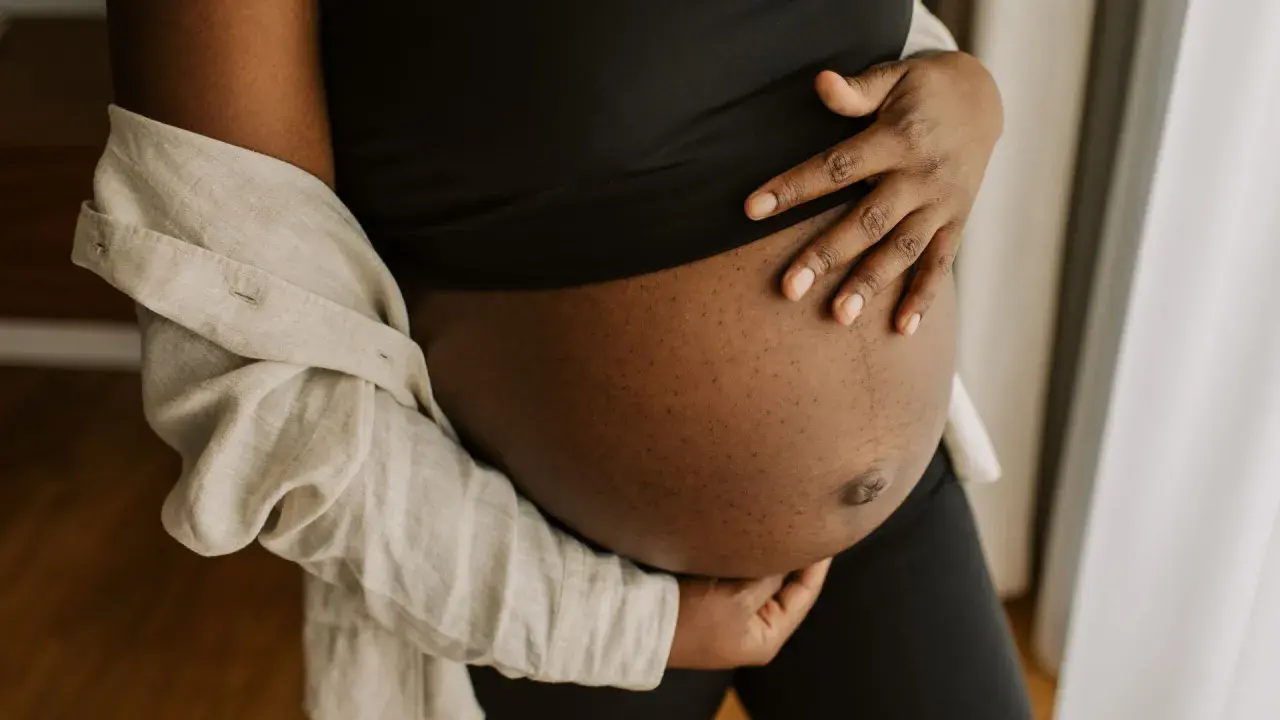 En gravid mage i tredje trimestern, varsamt omfamnad av händer. En ljus skjorta draperad över axeln.