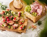 En härlig picknick med bruschetta toppad med jordgubbar och parmaskinka, färska vindruvor i korg och blommor.