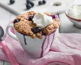 En nyttig mugcake med blåbär och en klick grädde, serverad i en vit mugg med lila droppar. En sked och en skål med grädde syns i bakgrunden.