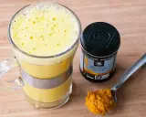 En gyllene dryck i ett glas, bredvid en burk ekologisk gurkmeja och en sked med kryddan. Perfekt för ditt nästa gurkmeja svartpeppar recept.