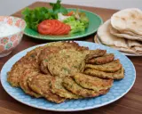 En stor tallrik med gyllene zeinas zucchinibiffar, serverade med pitabröd, en skål med vit sås och en sallad med tomat och gurka.