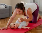 En mamma på knä på en röd yogamatta leker med sin bebis. Hon hjälper till med bebisens ben. En fin stund för post partum-träning.