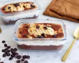 Två glasbehållare med nyttig chokladpudding, toppad med banan, nötter och chokladknappar. En gyllene sked ligger bredvid.