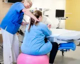 Barnmorska ger stöd till en kvinna i gravid vecka 36 som sitter på en rosa boll vid en förlossningssäng.