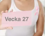 En person håller upp ett rosa kort med texten "Vecka 27". En rosa band pryder personens vita linne.