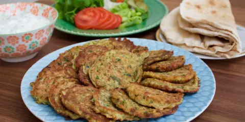 En stor tallrik med gyllene zeinas zucchinibiffar, serverade med pitabröd, en skål med vit sås och en sallad med tomat och gurka.