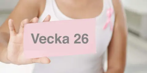 En person håller upp en rosa lapp med texten "Vecka 26". En rosa band-symbol syns på personens vita kläder.