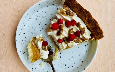 En bit glutenfri pumpapaj med vispgrädde, lingon och rostade pumpafrön på en prickig tallrik. En sked med en bit paj ligger bredvid.