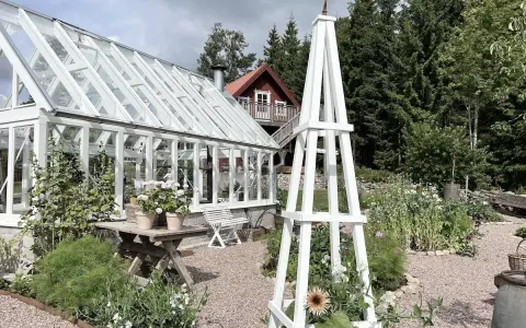 En vit rosentorn ritning står i en lummig trädgård med växthus och blommor.