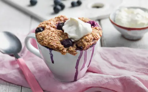 En nyttig mugcake med blåbär och en klick grädde, serverad i en vit mugg med lila droppar. En sked och en skål med grädde syns i bakgrunden.