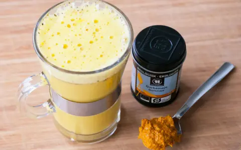 En gyllene dryck i ett glas, bredvid en burk ekologisk gurkmeja och en sked med kryddan. Perfekt för ditt nästa gurkmeja svartpeppar recept.