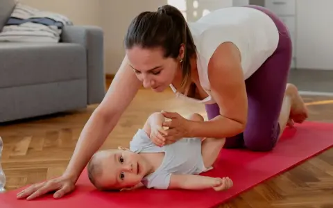 En mamma på knä på en röd yogamatta leker med sin bebis. Hon hjälper till med bebisens ben. En fin stund för post partum-träning.