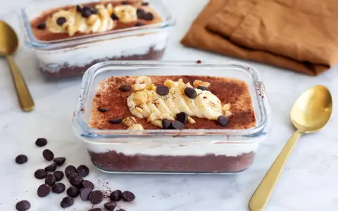 Två glasbehållare med nyttig chokladpudding, toppad med banan, nötter och chokladknappar. En gyllene sked ligger bredvid.