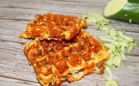 Gyldene zucchini våfflor, perfekt stekta, med riven zucchini och en hel zucchini i bakgrunden.