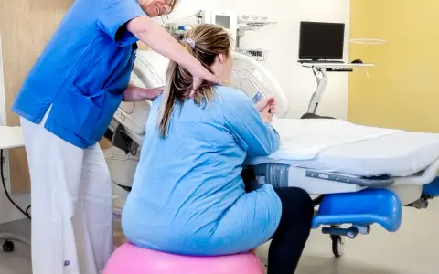 Barnmorska ger stöd till en kvinna i gravid vecka 36 som sitter på en rosa boll vid en förlossningssäng.