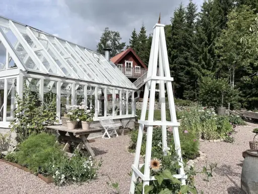 En vit rosentorn ritning står i en lummig trädgård med växthus och blommor.
