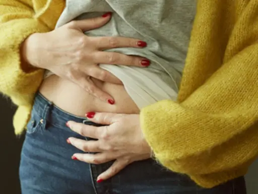 En kvinna med röda naglar håller om sin mage, klädd i en gul kofta och jeans. Kan man vara gravid trots mens?