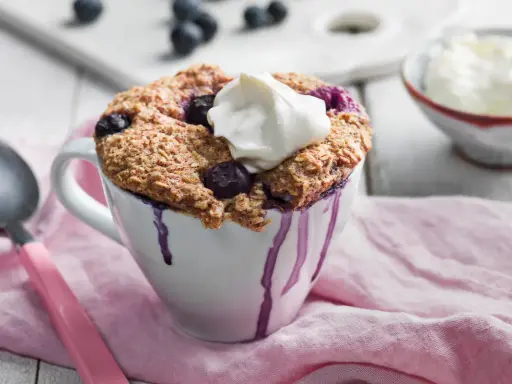 En nyttig mugcake med blåbär och en klick grädde, serverad i en vit mugg med lila droppar. En sked och en skål med grädde syns i bakgrunden.