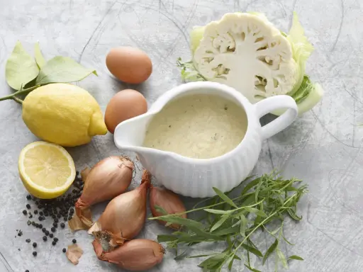 En skål med nyttig bearnaisesås omgiven av ingredienser som citron, ägg, schalottenlök, pepparkorn, dragon och blomkål.