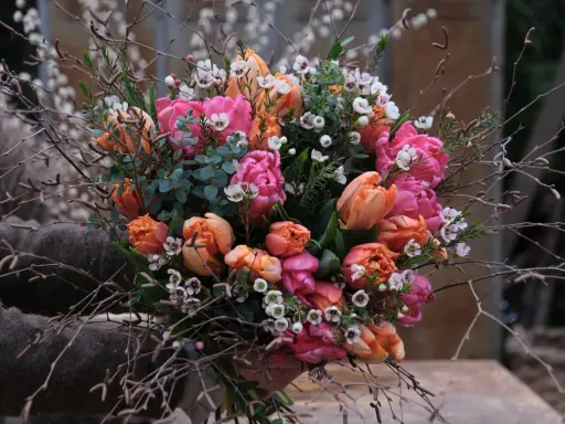En bukett med rosa och orange tulpaner, omgiven av kvistar och små vita blommor. Vatten droppar från de gröna bladen.