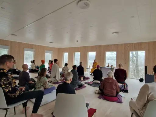 Människor sitter på kuddar och stolar under en meditation retreat i Sverige. En person i munkkläder leder sessionen.