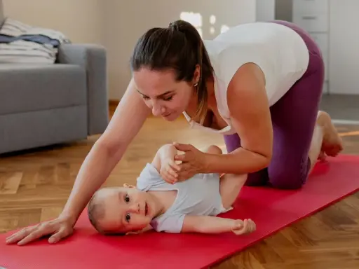 En mamma på knä på en röd yogamatta leker med sin bebis. Hon hjälper till med bebisens ben. En fin stund för post partum-träning.