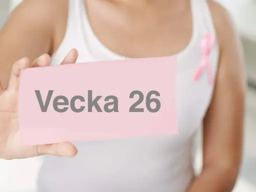 En person håller upp en rosa lapp med texten "Vecka 26". En rosa band-symbol syns på personens vita kläder.