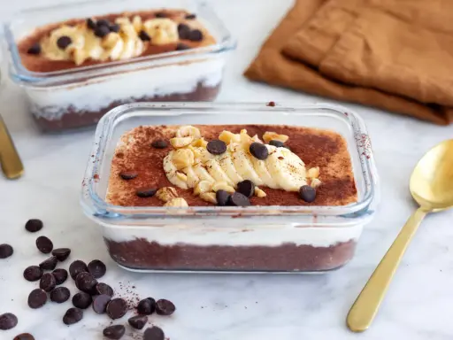 Två glasbehållare med nyttig chokladpudding, toppad med banan, nötter och chokladknappar. En gyllene sked ligger bredvid.