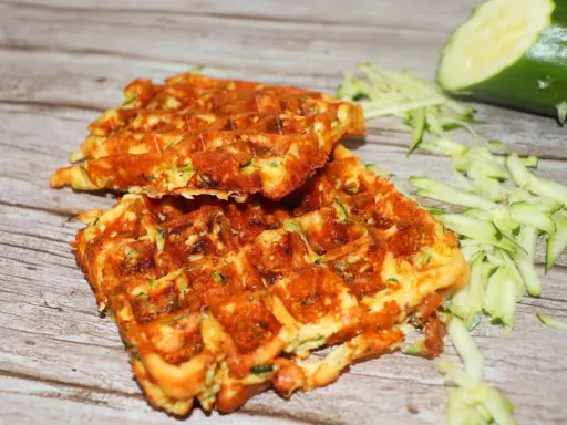 Gyldene zucchini våfflor, perfekt stekta, med riven zucchini och en hel zucchini i bakgrunden.