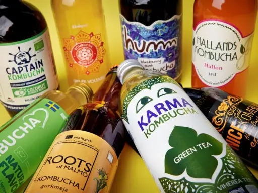 Flera flaskor kombucha, en nyttig och fermenterad dryck, ligger utspridda. Olika märken som Captain Kombucha, Karma Kombucha och Hallands Kombucha syns.