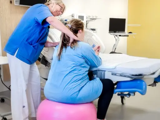 Barnmorska ger stöd till en kvinna i gravid vecka 36 som sitter på en rosa boll vid en förlossningssäng.
