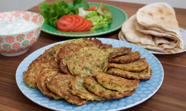 En stor tallrik med gyllene zeinas zucchinibiffar, serverade med pitabröd, en skål med vit sås och en sallad med tomat och gurka.