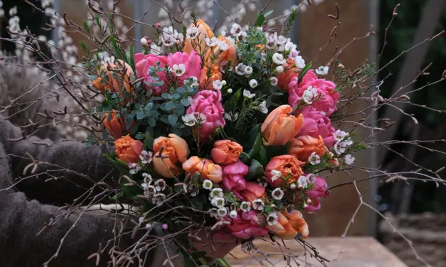 En bukett med rosa och orange tulpaner, omgiven av kvistar och små vita blommor. Vatten droppar från de gröna bladen.
