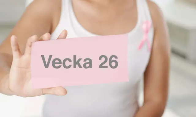 En person håller upp en rosa lapp med texten "Vecka 26". En rosa band-symbol syns på personens vita kläder.