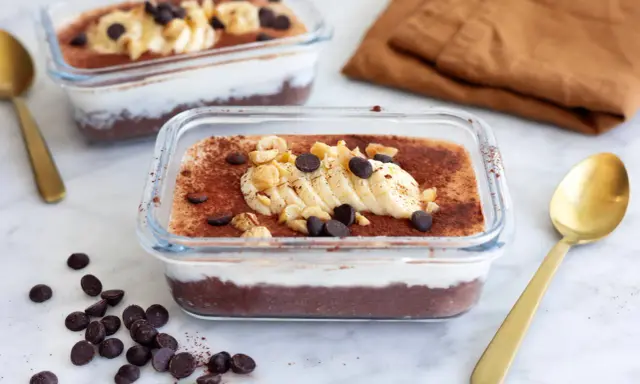 Två glasbehållare med nyttig chokladpudding, toppad med banan, nötter och chokladknappar. En gyllene sked ligger bredvid.
