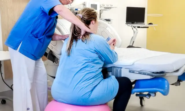 Barnmorska ger stöd till en kvinna i gravid vecka 36 som sitter på en rosa boll vid en förlossningssäng.