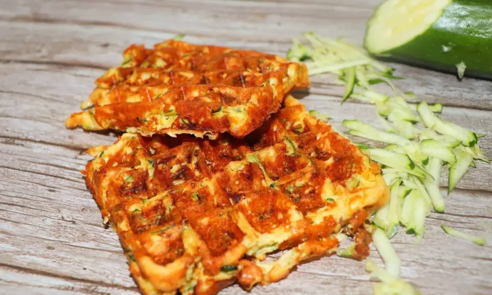 Gyldene zucchini våfflor, perfekt stekta, med riven zucchini och en hel zucchini i bakgrunden.