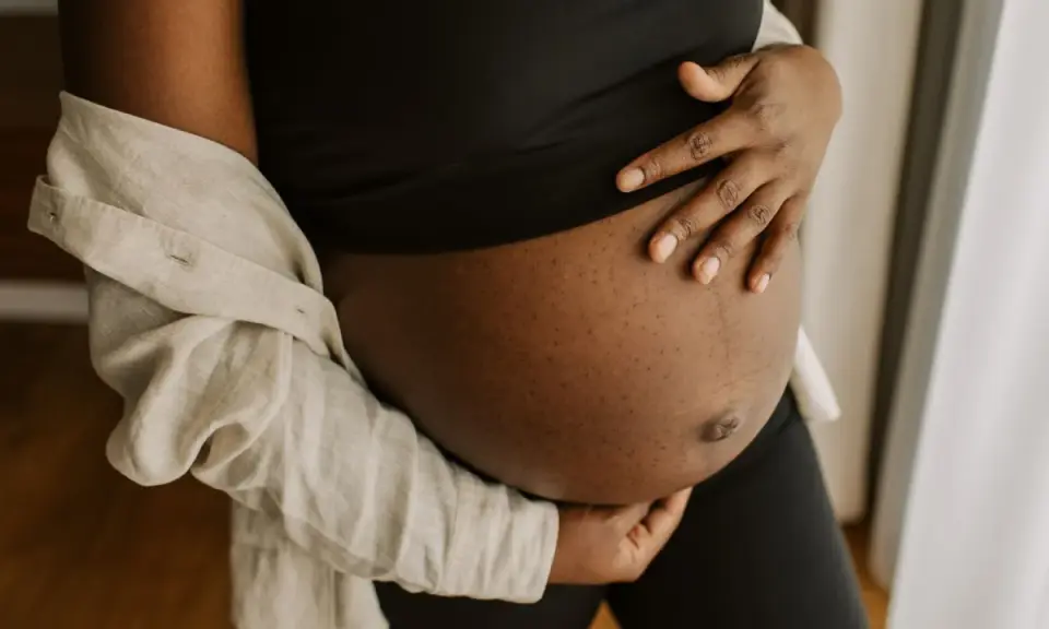 En gravid mage i tredje trimestern, varsamt omfamnad av händer. En ljus skjorta draperad över axeln.