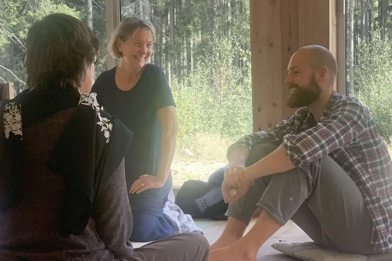 Tre personer ler och samtalar under en meditation retreat sverige. En kvinna st&aring;r, medan tv&aring; sitter p&aring; golvet.